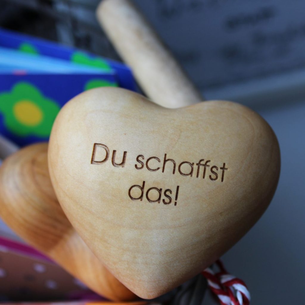 Holzherz mit Gravur „Du schaffst das!“ vor buntem Hintergrund